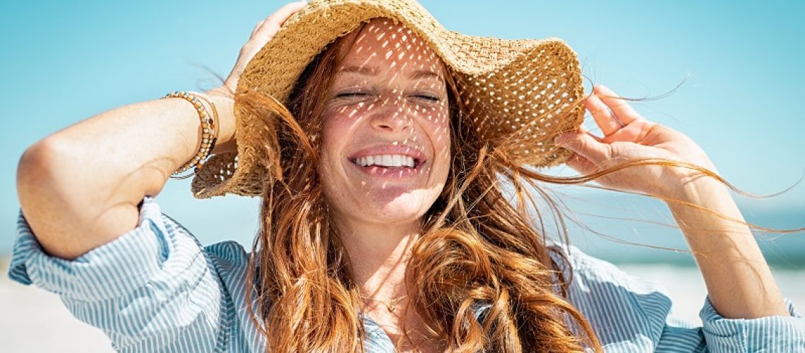Closeup face of mature woman wearing straw hat enjoying the sun at beach. Happy young woman smiling during summer vacation at sea. Portrait of beautiful lady relaxing at beach while holding large brim for the wind.